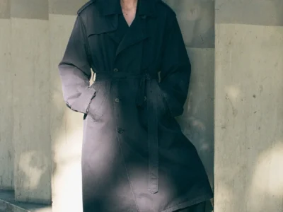 Model standing beside concrete columns wearing an oversized dark ssstein trench coat with a belted waist and relaxed black trousers in soft natural light