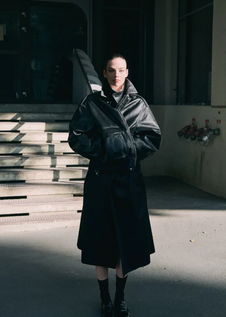 Model standing in dramatic sunlight wearing an oversized black ssstein leather jacket with sculptural sleeves and dark tailored layers against a stark architectural backdrop