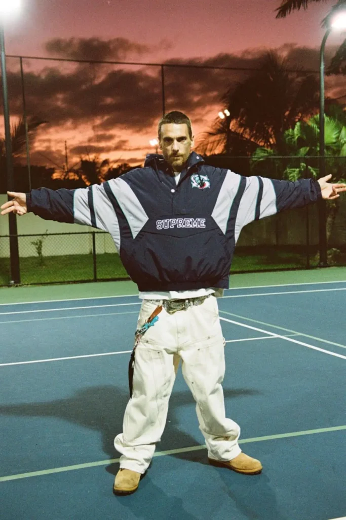 A moody campaign image from the Supreme x SALEM Spring/Summer 2026 collaboration showing a model standing on a tennis court at dusk beneath dramatic sunset skies and palm trees. The look centers around an oversized navy half-zip stadium jacket with gray paneling, SALEM-inspired graphics, and bold Supreme branding across the chest, paired with loose white pants and tan suede boots. The cinematic lighting and isolated setting reinforce the capsule’s dark, atmospheric identity rooted in underground music culture and late-night streetwear aesthetics