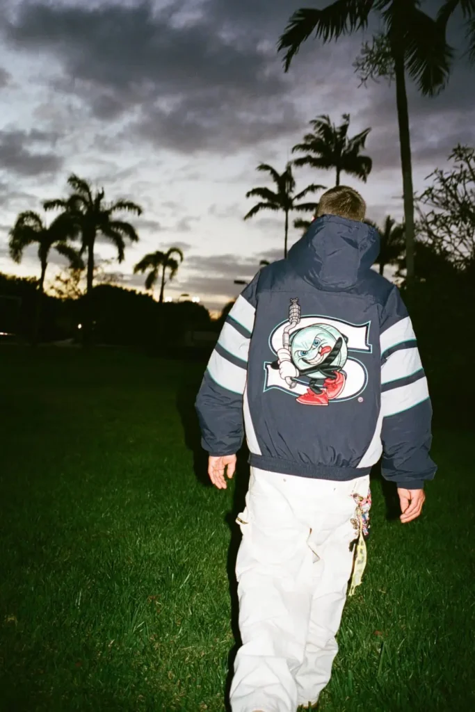 A nighttime campaign image from the Supreme x SALEM SS26 collaboration showing a model walking across a dark palm-lined lawn at dusk. The oversized navy stadium jacket features bold striped sleeves and a large graphic mascot emblem across the back, blending retro sportswear energy with SALEM’s surreal underground aesthetic. Flash photography and stormy evening skies give the image a cinematic, dreamlike atmosphere
