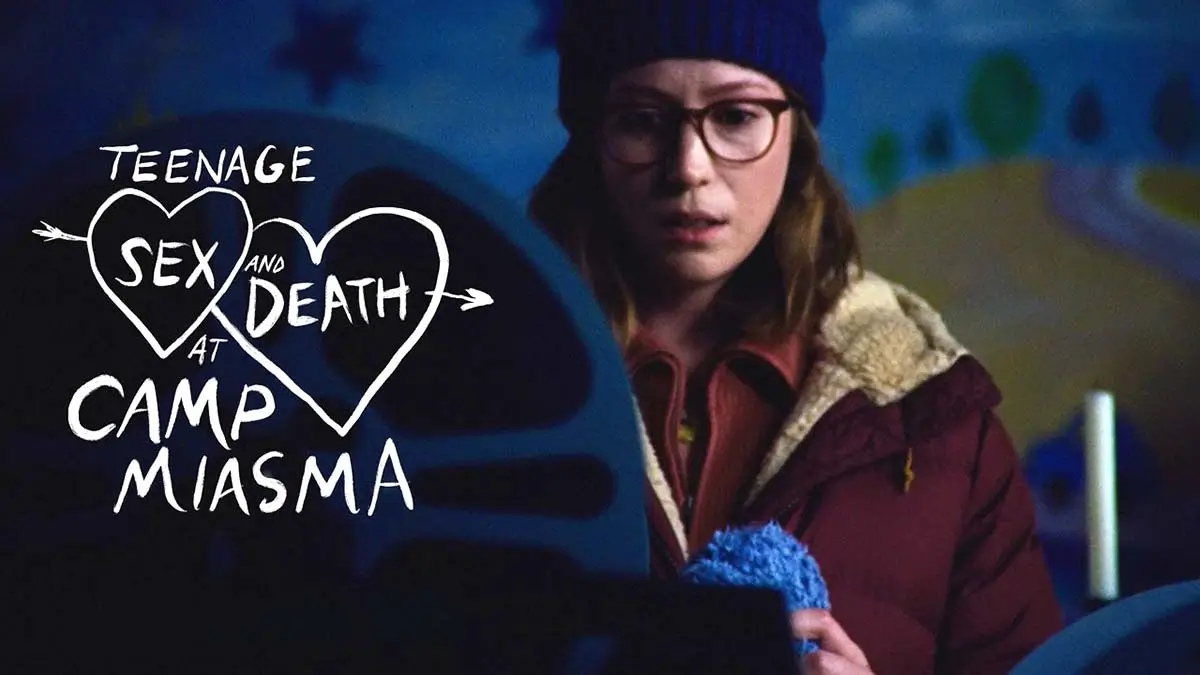 A frightened young woman in glasses and a knit beanie stares into the darkness beside the hand-drawn Teenage Sex and Death at Camp Miasma title card, blending indie coming-of-age aesthetics with eerie slasher tension. The image pairs soft analog textures and intimate character framing with ominous blue lighting, evoking the nostalgic VHS horror atmosphere central to Jane Schoenbrun’s Cannes breakout film