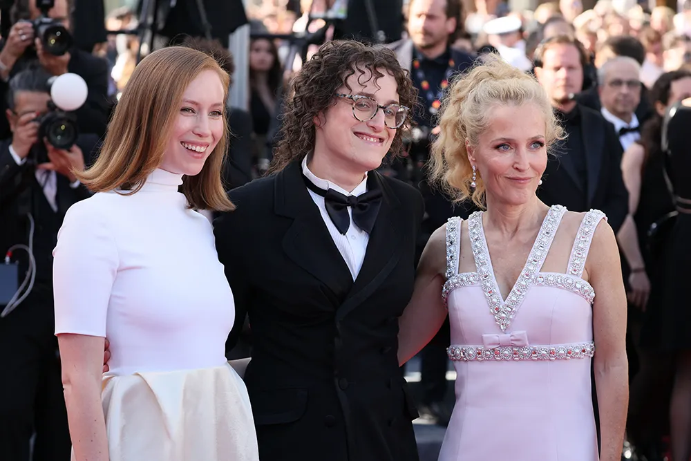 Jane Schoenbrun, Hannah Einbinder, and Gillian Anderson pose together on the Cannes Film Festival red carpet, presenting a striking mix of classic glamour and contemporary queer cinema cool. The image captures the trio celebrating Teenage Sex and Death at Camp Miasma amid flashing cameras, formal tailoring, pastel couture detailing, and the charged atmosphere surrounding one of Cannes 2026’s breakout films