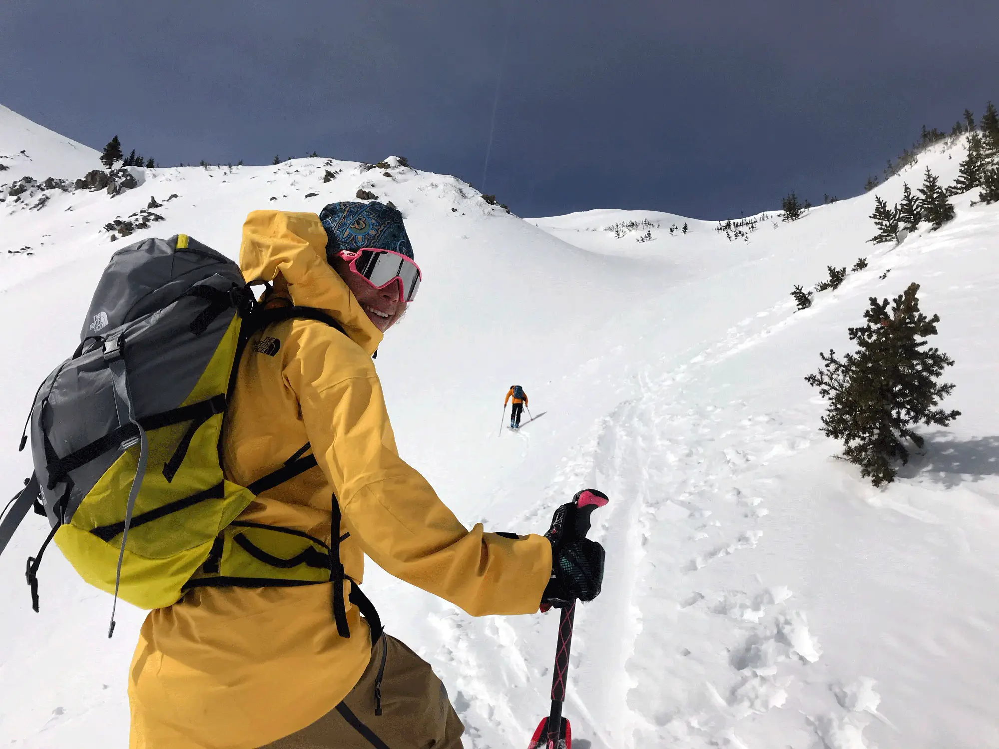 Backcountry skier in a yellow The North Face shell jacket ascending a snow-covered alpine ridge with touring gear and a large expedition backpack under dramatic mountain skies