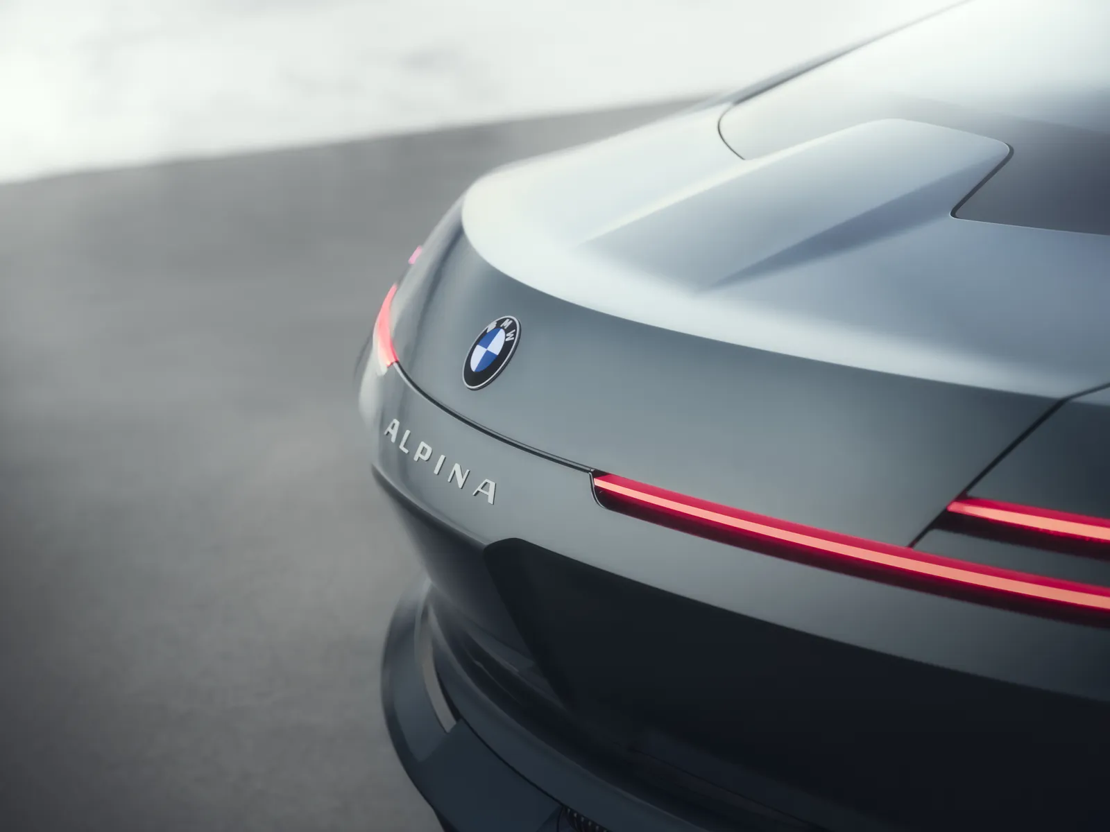 A minimalist rear-quarter view of the Vision BMW ALPINA highlights the concept’s sculptural tail design and razor-thin red light signature stretching seamlessly across the bodywork. The softly illuminated ALPINA lettering sits beneath the BMW roundel, reinforcing the vehicle’s understated branding philosophy, while the integrated ducktail spoiler and flowing rear surfaces emphasize aerodynamic elegance over overt aggression. Wrapped in muted metallic paint and surrounded by a foggy monochromatic backdrop, the composition captures the grand tourer’s calm, architectural presence—more luxury object than conventional performance coupe