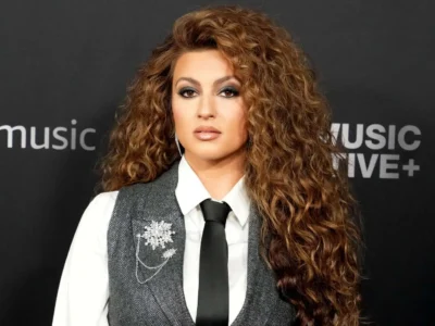 Tori Kelly delivers a polished, menswear-inspired red-carpet look with a crisp white shirt, black tie, and textured charcoal vest accented by jeweled brooch details. Her voluminous curls soften the structured tailoring, creating a balance between sophistication and glamour. Paired with smoky makeup and a dark backdrop, the overall styling feels elegant, confident, and reflective of her more mature artistic era