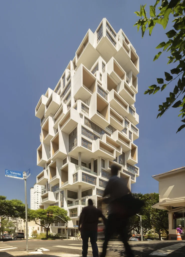 A striking contemporary high-rise composed of stacked cantilevered concrete volumes rises dramatically above a city intersection under a clear blue sky. The sculptural residential tower features deep recessed balconies, geometric protrusions, and expansive bronze-framed glazing, while blurred pedestrians crossing in the foreground introduce motion, scale, and an active urban atmosphere against the building’s monumental architectural presence