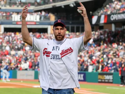 Travis Kelce stands on the field at Progressive Field wearing a Cleveland Guardians jersey and cap, raising both arms while holding a baseball during a ceremonial appearance before a packed crowd