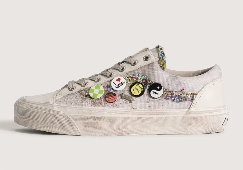 Side-profile product shot of the Vans Old Skool 36 “Souvenir” sneaker in an oatmeal colorway against a light neutral background. The low-top shoe features a textured canvas upper with frayed multicolor woven detailing replacing the traditional Vans side stripe, creating a handcrafted patchwork effect. Several decorative pin badges—including checkerboard, smiley face, yin-yang, and “I ♥ Vans” graphics—are attached across the midfoot. Distressed foxing, tonal laces, and lightly weathered midsoles give the shoe a worn-in vintage aesthetic inspired by DIY skate and souvenir culture
