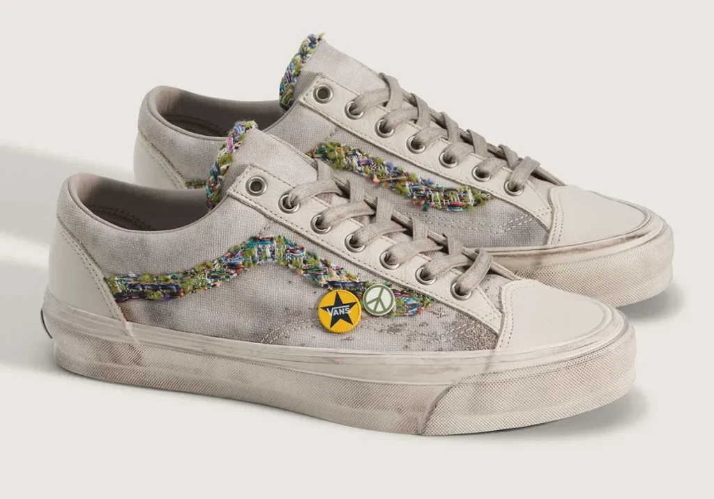 ngled product shot of the Vans Old Skool 36 “Souvenir” shoe in an oatmeal colorway, displayed as a pair against a soft neutral background. The shoe feature textured canvas uppers with distressed detailing, off-white leather overlays, and frayed multicolor woven jazz stripes that create a handcrafted patchwork appearance. Tonal laces and metallic eyelets maintain the vintage skate silhouette, while decorative peace-sign and Vans star pins attached near the side panels reinforce the shoe’s DIY customization theme. Aged foxing and lightly weathered midsoles complete the worn-in aesthetic inspired by nostalgia, skate culture, and collected memorabilia
