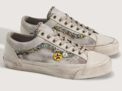 ngled product shot of the Vans Old Skool 36 “Souvenir” shoe in an oatmeal colorway, displayed as a pair against a soft neutral background. The shoe feature textured canvas uppers with distressed detailing, off-white leather overlays, and frayed multicolor woven jazz stripes that create a handcrafted patchwork appearance. Tonal laces and metallic eyelets maintain the vintage skate silhouette, while decorative peace-sign and Vans star pins attached near the side panels reinforce the shoe’s DIY customization theme. Aged foxing and lightly weathered midsoles complete the worn-in aesthetic inspired by nostalgia, skate culture, and collected memorabilia