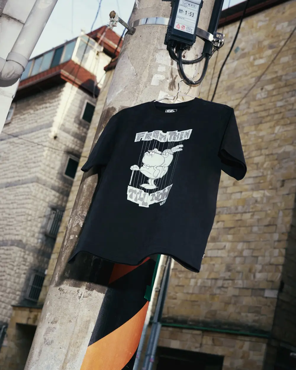 Editorial product image from the Vans x SOON.EASY collection featuring a black graphic T-shirt suspended against an urban utility pole backdrop. The tee showcases distorted typography and a playful cartoon-style illustration rendered in SOON.EASY’s signature satirical visual language. Surrounded by weathered architecture, exposed cables, and street textures, the campaign styling reinforces the collection’s raw, rebellious blend of contemporary art and streetwear culture