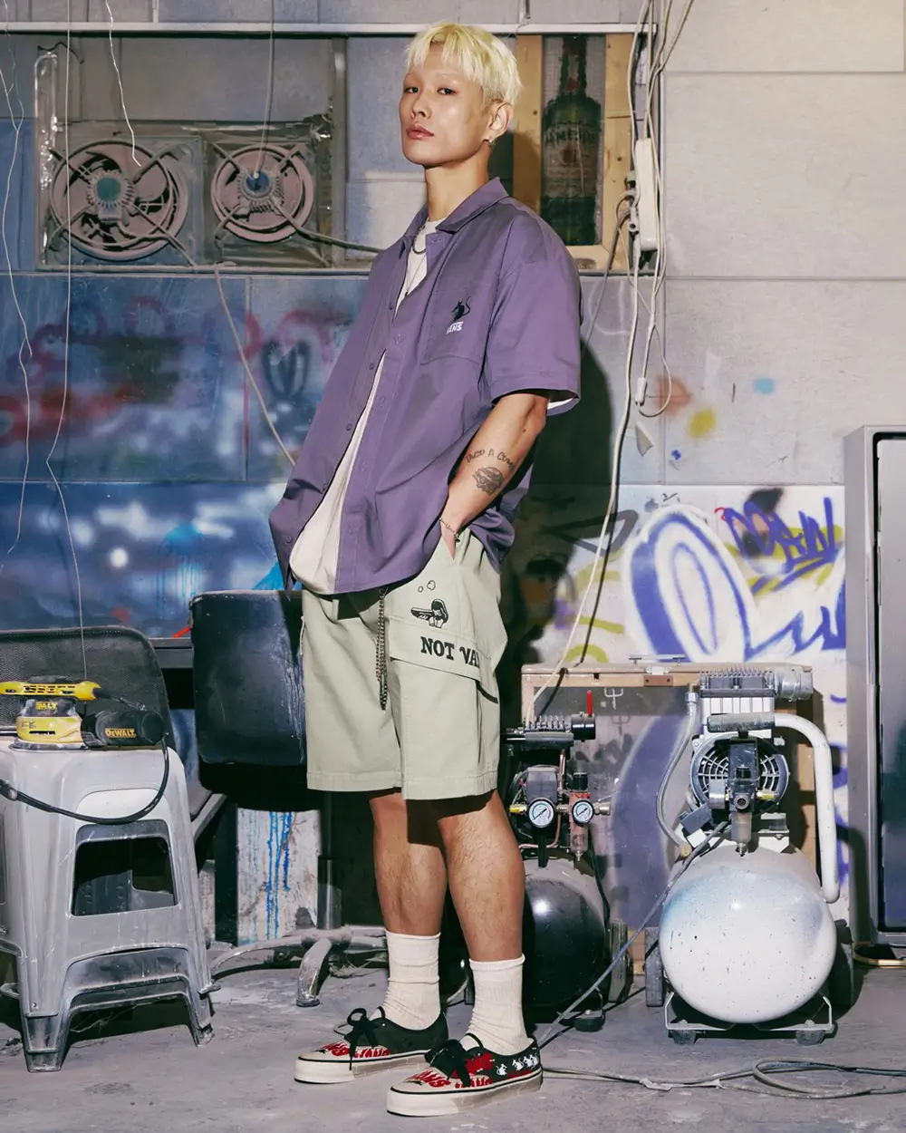 editorial campaign image featuring a model wearing pieces from the Vans x SOON.EASY collection inside an industrial, graffiti-marked studio space. The look pairs an oversized muted purple short-sleeve button-up with graphic cargo shorts, white crew socks, and the illustrated Vans x SOON.EASY Premium LX Authentic sneakers. Surrounded by exposed wires, workshop equipment, and spray-painted surfaces, the setting amplifies the collection’s rebellious, art-meets-streetwear identity and raw underground atmosphere