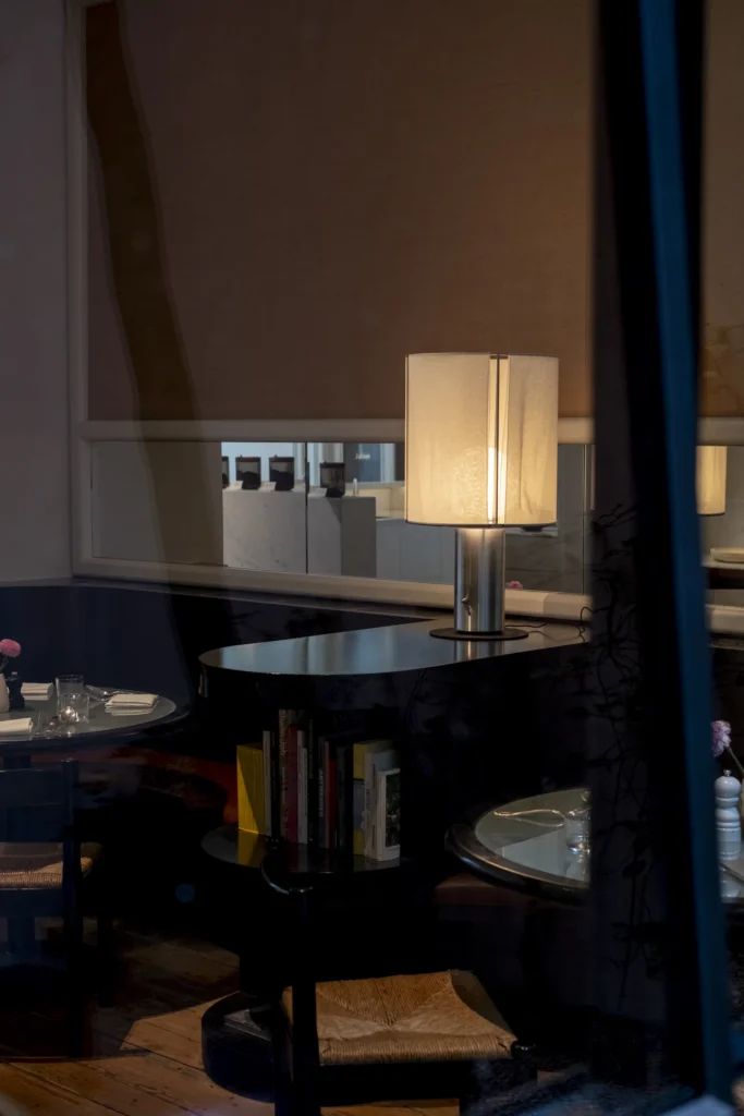 A softly lit restaurant interior is viewed through reflective glass, creating a layered, cinematic atmosphere of warm shadows and muted elegance. A modern table lamp glows beside curved black shelving filled with books, while intimate dining tables with woven chairs and neatly arranged place settings suggest a quiet late-evening ambiance