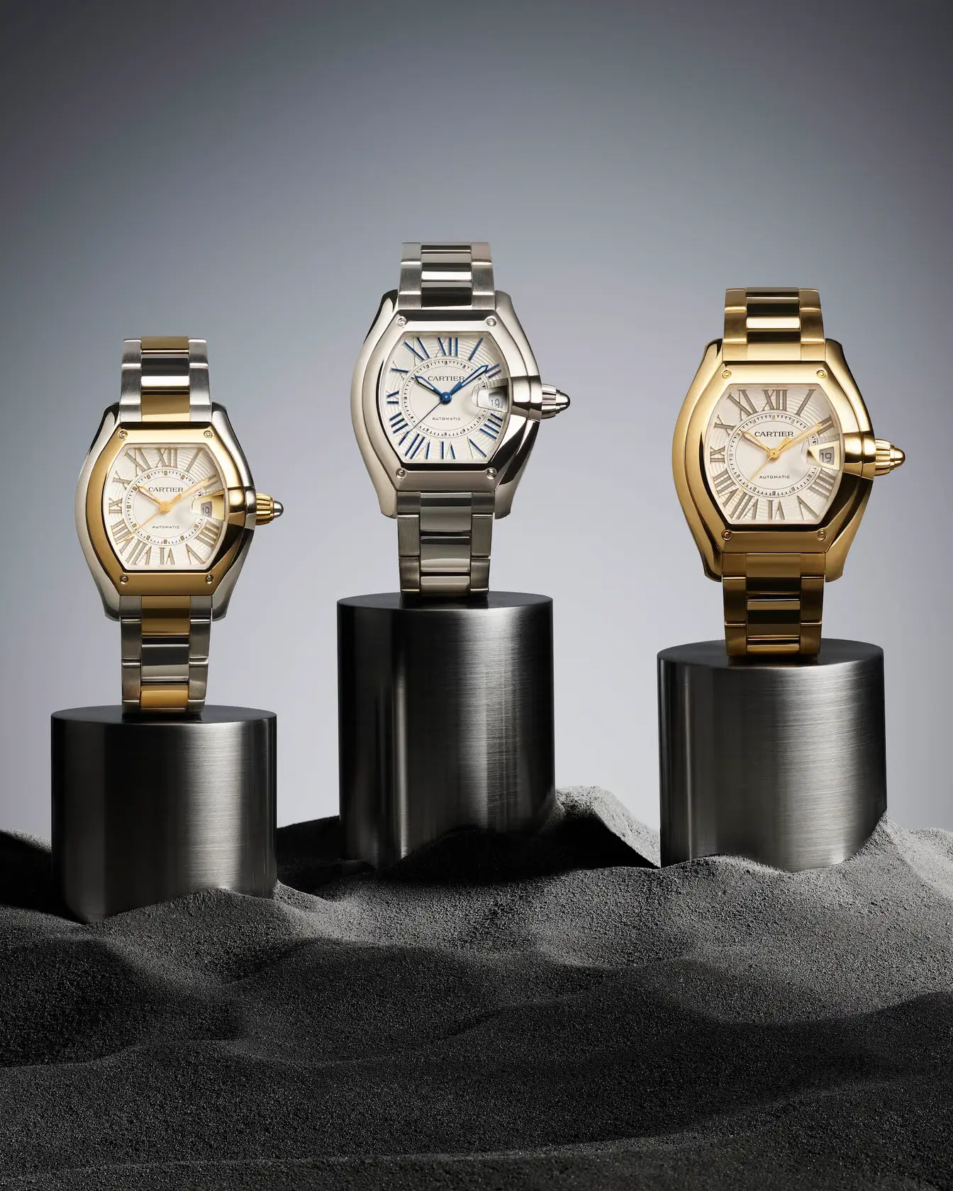 Three Cartier Roadster 2026 watches displayed on cylindrical podiums set in textured dark sand, showcasing stainless steel, two-tone steel and yellow gold, and full yellow gold executions. Each model features the collection’s signature tonneau-shaped case, Roman numeral dial, integrated bracelet, and magnified date window in a clean studio setting