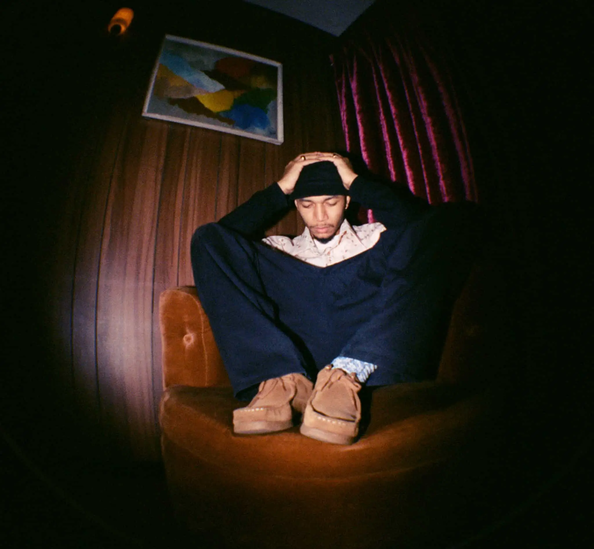 A moody portrait of a seated musician in a dimly lit, wood-paneled room, captured with a wide-angle, slightly distorted perspective. The subject sits curled into a vintage brown armchair with hands resting on their head, wearing layered dark clothing, a black cap, patterned socks, and soft suede shoes. Deep burgundy curtains and an abstract framed artwork create a cinematic backdrop, while the low lighting and intimate posture evoke introspection, late-night reflection, and a quietly contemplative atmosphe