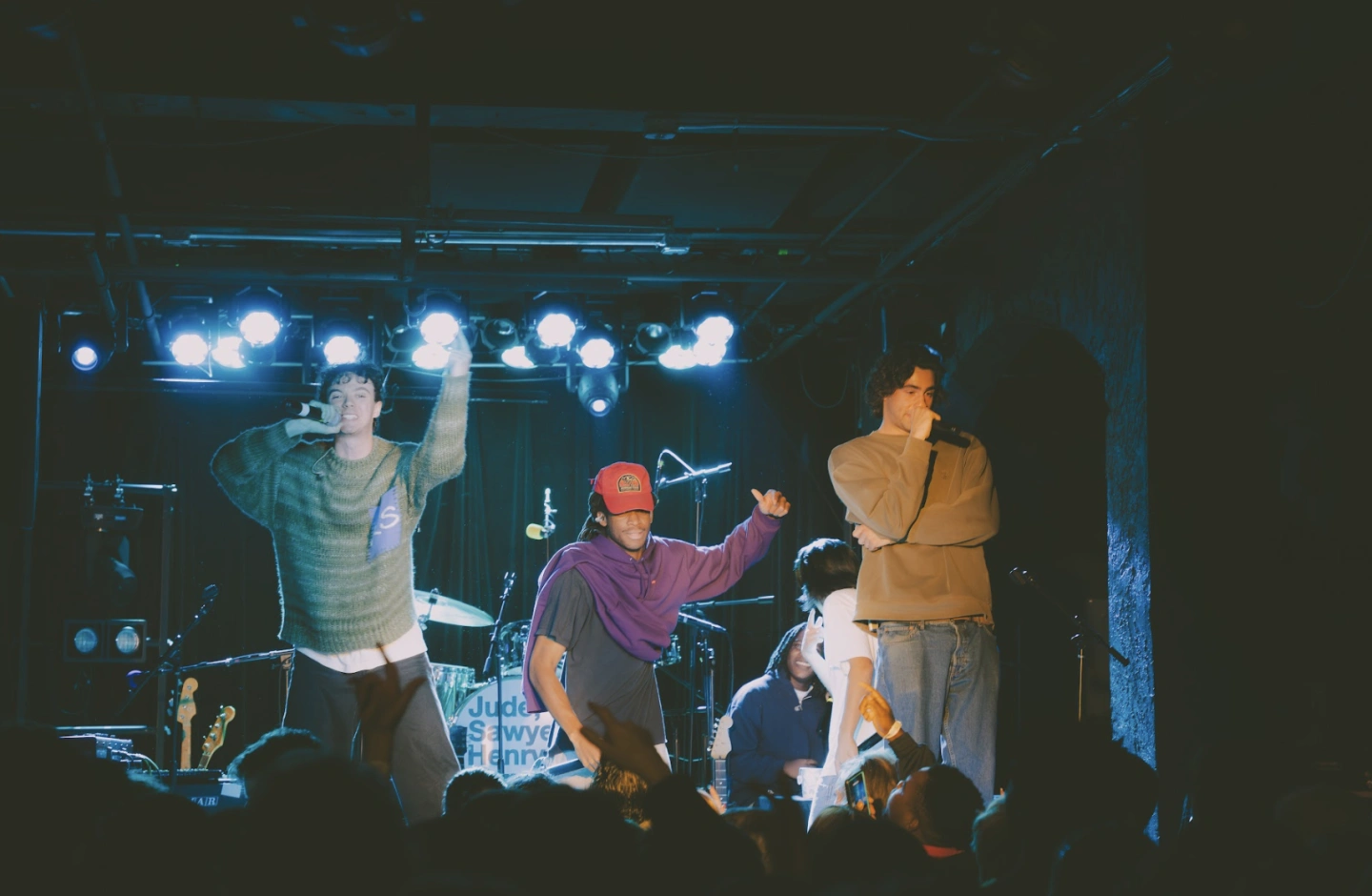 Performance photo of WHATMORE on stage during an intimate live set, with members rapping and moving through bright blue concert lighting while the crowd raises hands from the front row