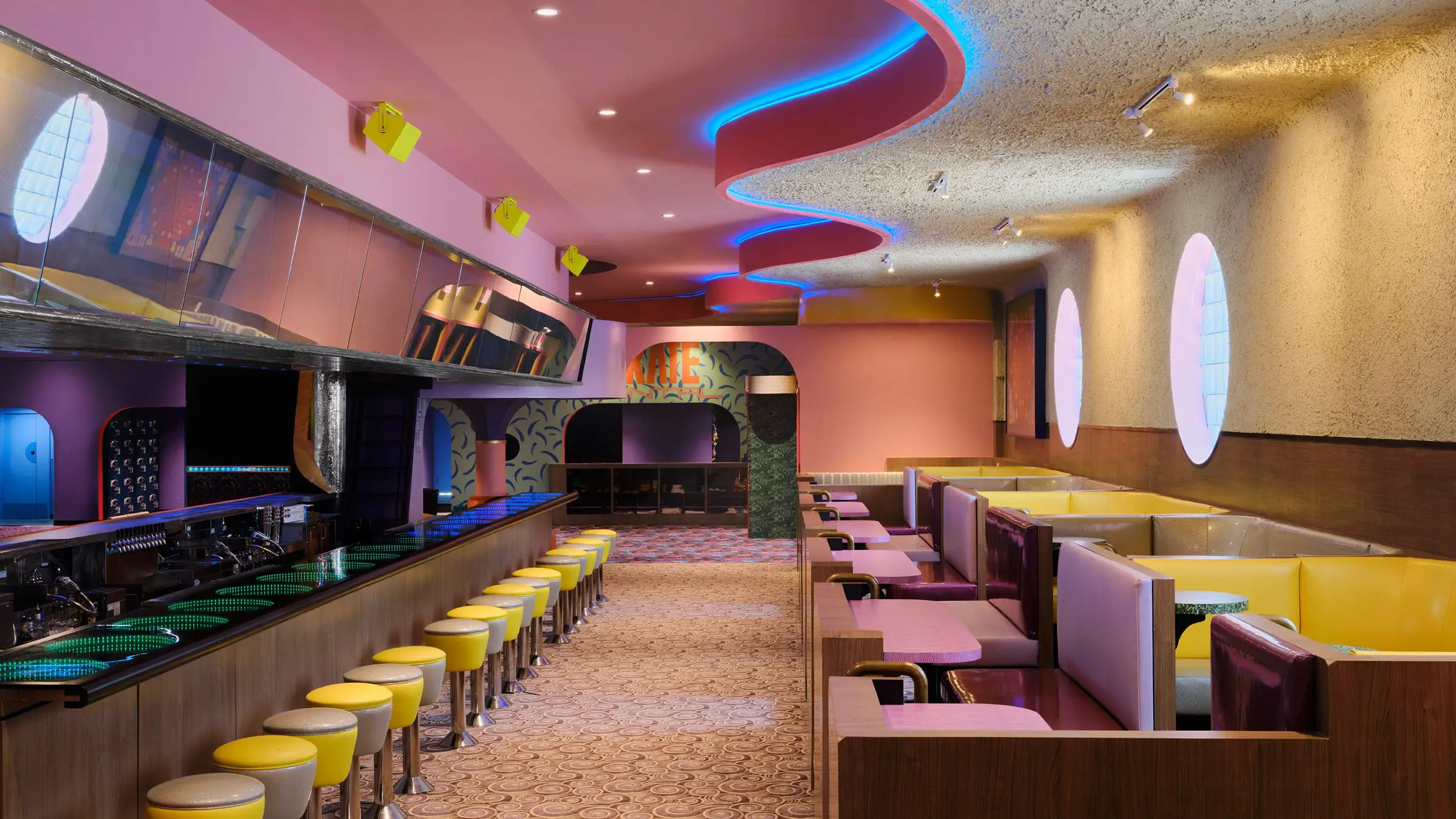 Wide interior view of a colorful retro-futurist lounge and bar space featuring pastel pink ceilings, curved architectural details, glowing blue accent lighting, and circular porthole-style windows. A long bar lined with yellow stools runs along one side while geometric booths in cream, yellow, and burgundy create intimate seating areas, blending roller-rink nostalgia with contemporary hospitality design