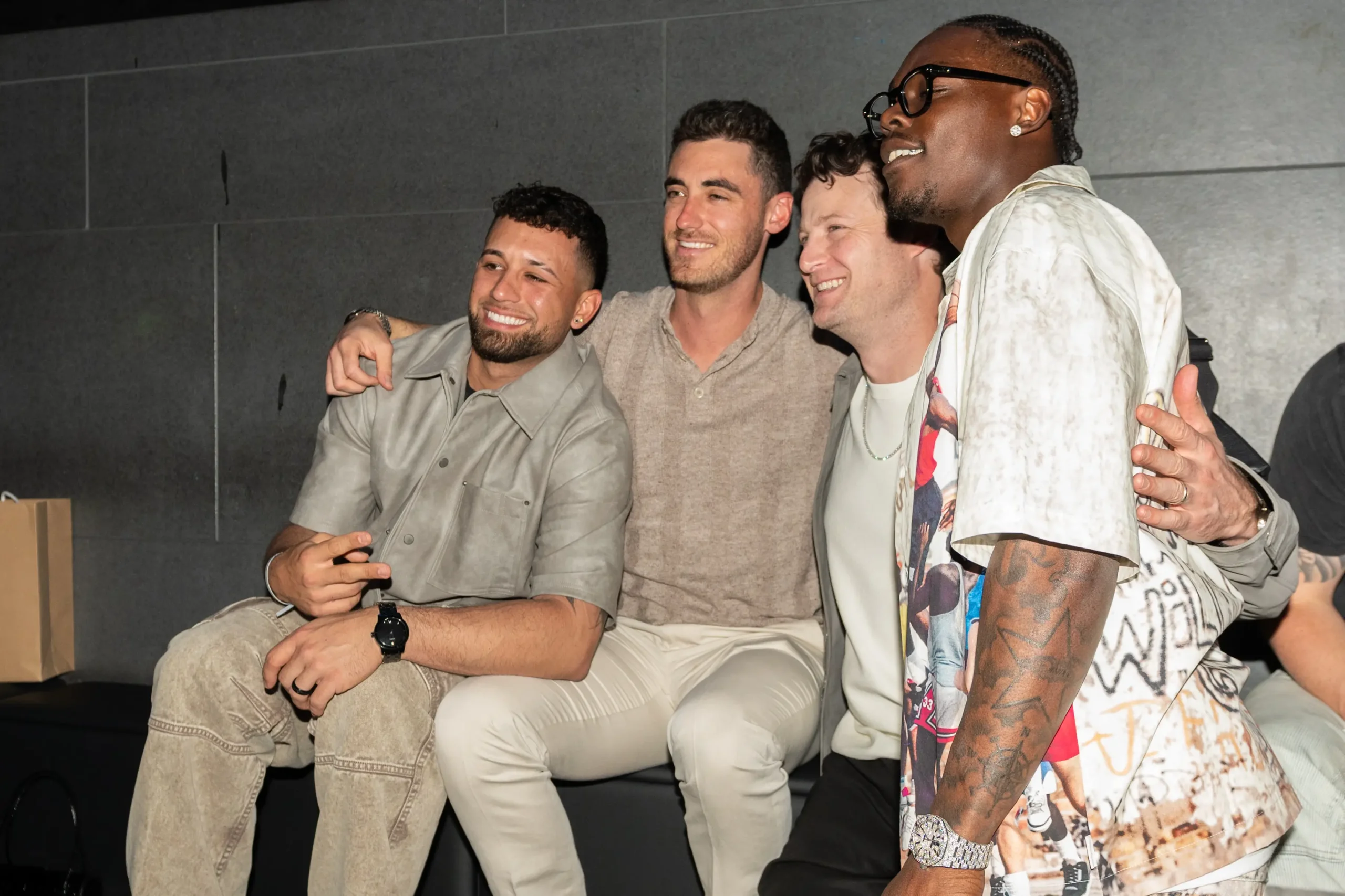 Jazz Chisholm Jr. poses with friends during a relaxed nighttime gathering, wearing a graphic oversized shirt, black trousers, luxury watch, and tinted glasses while seated against a dark modern interior backdrop