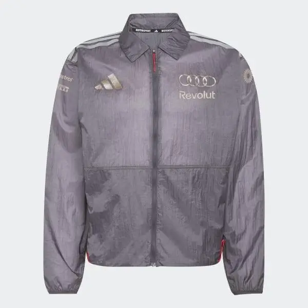 Grey lightweight zip-up jacket with a slightly crinkled, technical finish and a clean, athletic silhouette. The design features a full-length front zipper, elasticated cuffs, and a relaxed hem for functional comfort. The chest is detailed with multiple sponsor-style logos, including the Audi rings, “Revolut,” and an Adidas mark, alongside subtle additional branding on the sleeves. Tonal striping runs along the shoulders, reinforcing its performance-inspired identity. The muted grey palette keeps the look understated while the layered logos evoke a modern Formula 1 teamwear aesthetic translated into everyday outerwear