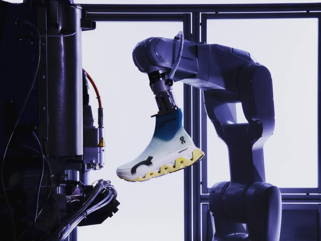 Robotic arm in a controlled lab setting holding a completed On LightSpray running shoe, with the seamless, sock-like upper and sculpted sole clearly visible against a bright, backlit industrial backdrop