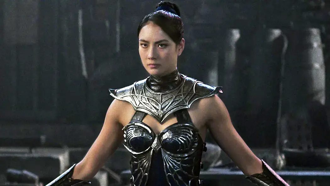 Character still of a warrior woman in dark ceremonial armor standing in a shadowy fantasy environment. The costume features sculpted metallic detailing across the chest and shoulders with layered black textures, while her hair is styled in a high knot. Her focused expression and upright stance convey authority, discipline, and calm intensity, emphasizing a regal yet battle-ready presence