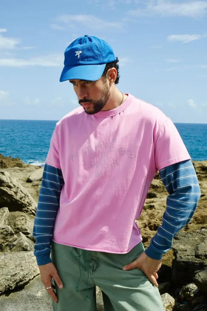 A man poses along a rocky oceanfront wearing layered casual separates built around soft pastel tones. The look combines a bright blue cap, a faded pink oversized T-shirt over a blue striped long-sleeve layer, and muted green bottoms, creating a relaxed coastal palette that feels sun-washed, playful, and effortlessly styled