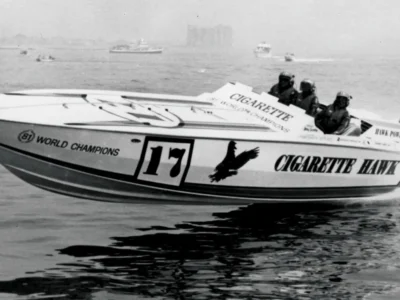 Black-and-white archival photograph of the Cigarette Hawk offshore powerboat, race number 17, skimming across calm water during a high-speed competition. Three helmeted crew members sit in the open cockpit as the boat, marked with "World Champions" and large Cigarette Hawk branding, lifts slightly above the surface, with distant boats and a hazy shoreline view in the background