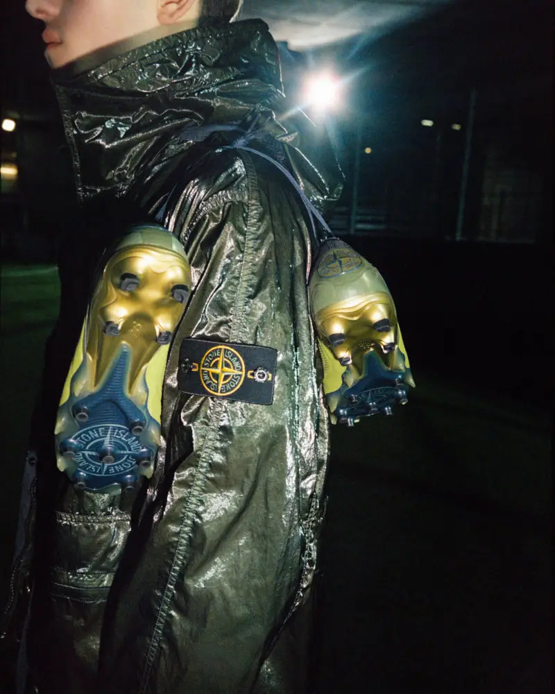 Close-up campaign image of a model wearing a metallic Stone Island technical jacket with New Balance football boots attached to the garment, highlighting the collection’s blend of sport and fashion