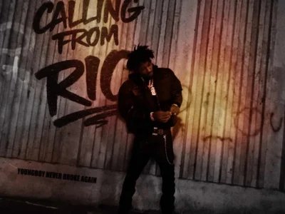 YoungBoy Never Broke Again stands in front of a weathered wooden wall beneath bold graffiti reading "Calling From Rio" on the cover art for the single. The artist wears a dark jacket, black pants with a chain accessory, layered diamond jewelry, and looks down while adjusting his watch. Warm orange lighting creates a gritty nighttime atmosphere against the industrial backdrop. "YoungBoy Never Broke Again" appears in the lower left corner alongside the BYNOAH credit, while a black-and-white Parental Advisory Explicit Content label sits in the bottom right