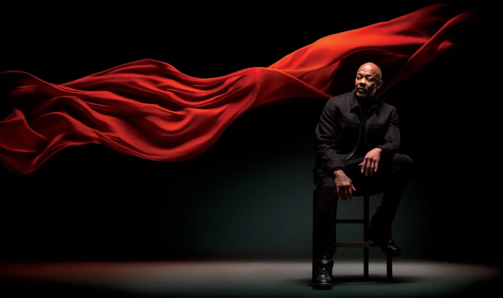 A seated Dr Dre dressed in black is framed by a sweeping wave of flowing red fabric against a dark studio background, creating a dramatic and cinematic portrait