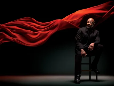 A seated Dr Dre dressed in black is framed by a sweeping wave of flowing red fabric against a dark studio background, creating a dramatic and cinematic portrait