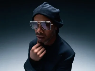 Ronaldinho poses in an oversized black suit, beret, and statement sunglasses while looking up toward the camera in a dramatic studio set