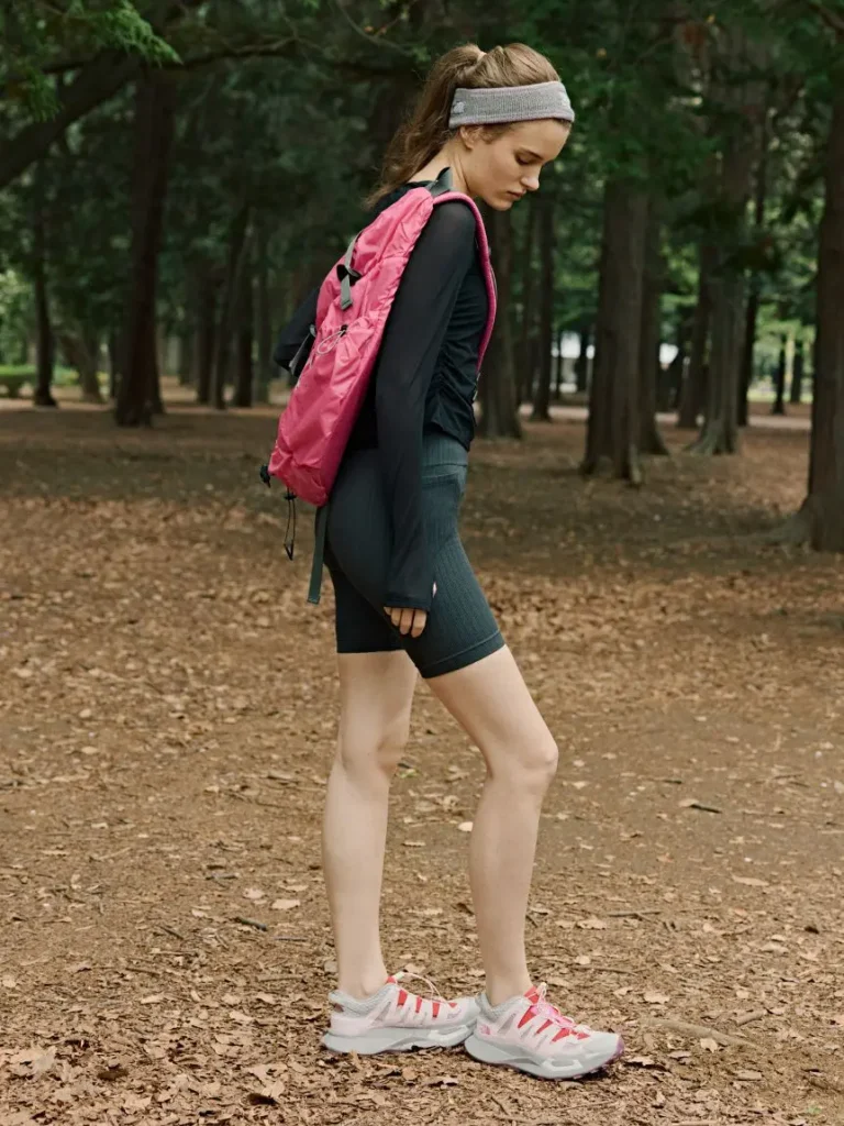 Outdoor lifestyle image of a model wearing The North Face VECTIV Breeze trail shoes with activewear and a pink backpack in a wooded set