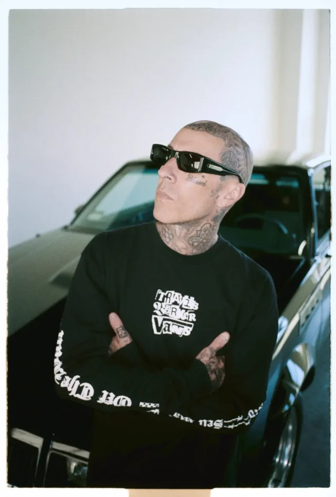 Portrait of a bald man with a thick mustache wearing a white Travis Barker x Vans T-shirt, standing against a chain-link fence with a playful expression