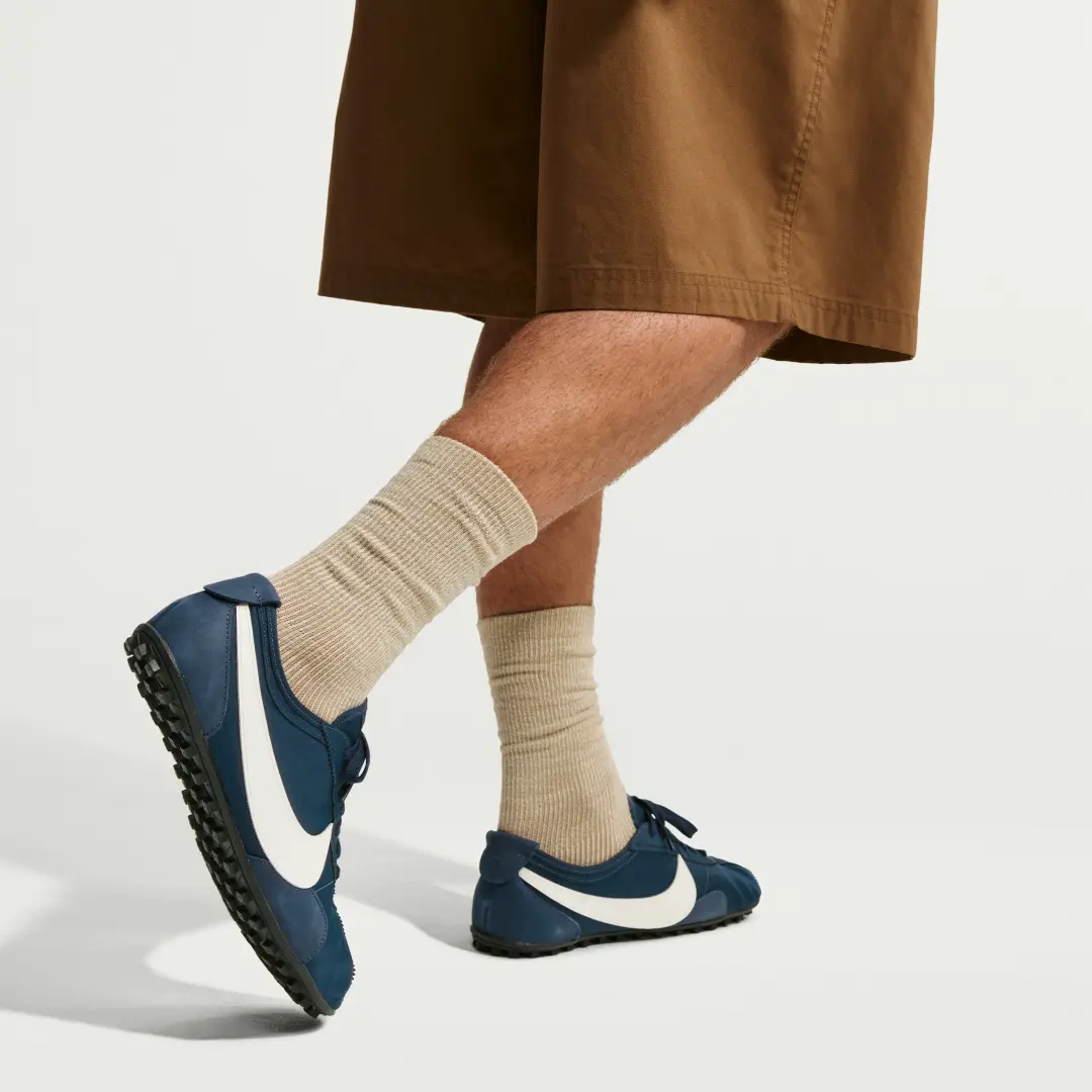 Lifestyle shot of a model wearing navy blue Nike Cortez-style sneakers with oversized white Swooshes, paired with beige ribbed crew socks and cropped brown trousers, highlighting the shoe’s sleek profile and rugged black lug outsole in a casual everyday set