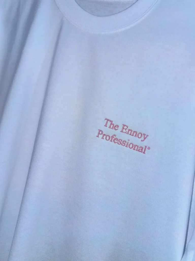 Close-up of a white crewneck T-shirt featuring minimalist chest embroidery reading "The Ennoy Professional®" in soft pink thread. The premium heavyweight cotton fabric, clean neckline, and understated branding emphasize the label's signature minimalist aesthetic