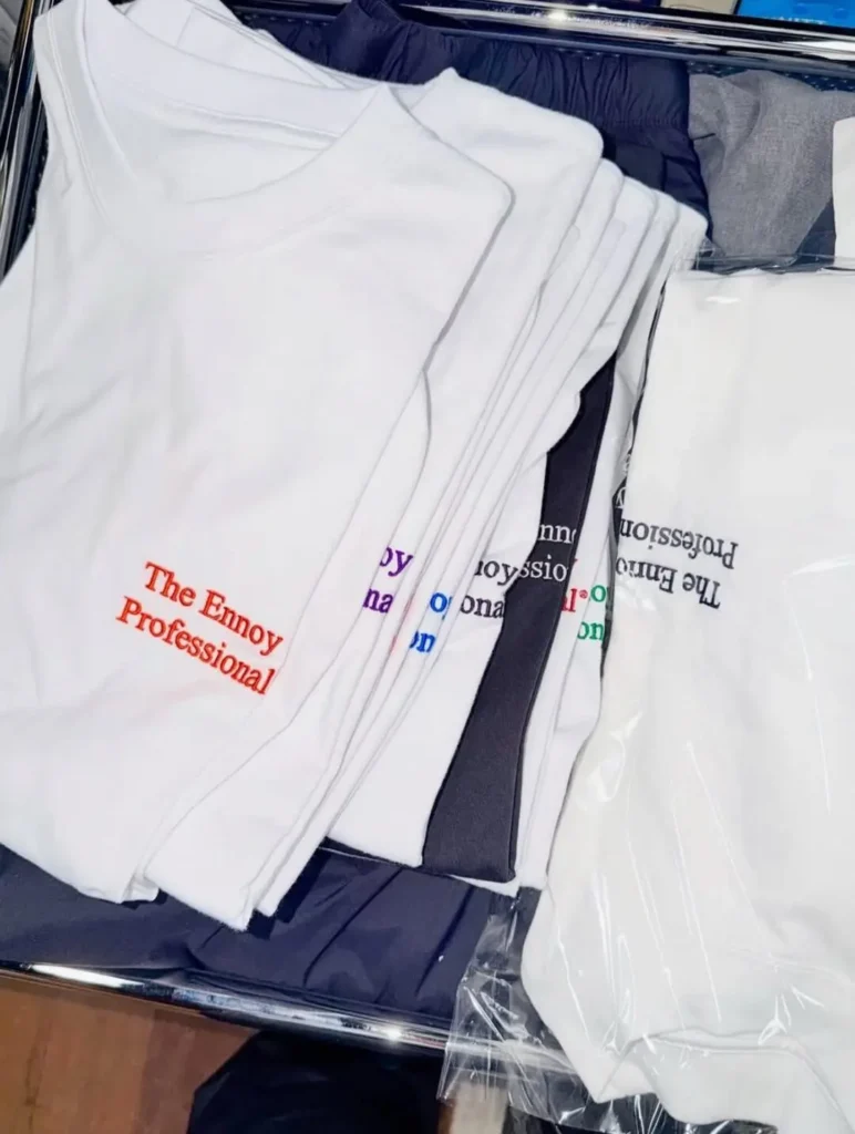 A stack of folded The Ennoy Professional® T-shirts displayed inside a storage bin, showcasing embroidered logo color variations including red, purple, blue, black, green, and pink. White tees are paired with black garments, highlighting the collection's simple, color-focused brand