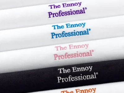 A neatly arranged lineup of folded The Ennoy Professional® T-shirts displaying embroidered chest logos in multiple colors, including red, black, purple, blue, pink, white, orange, and green. The clean presentation highlights the brand's understated design and range of embroidery options