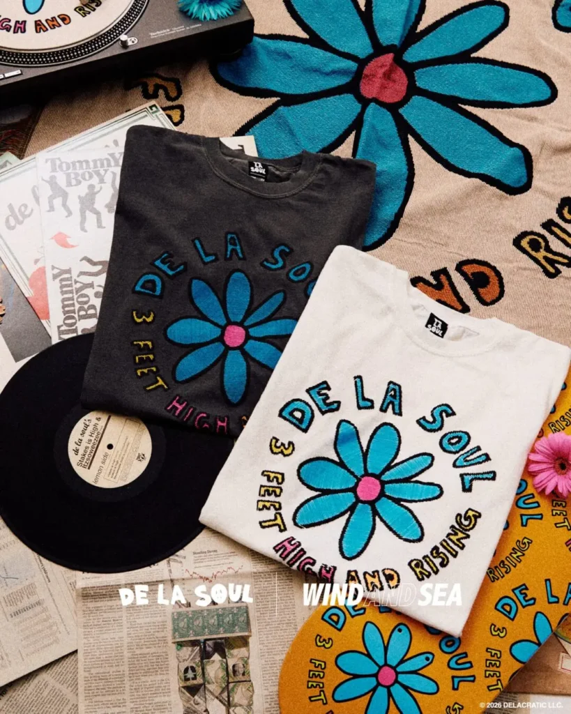 Black and white De La Soul x WIND AND SEA T-shirts featuring the group's signature blue flower logo, arranged on a retro-inspired backdrop with vinyl records, printed ephemera, and collectible memorabilia