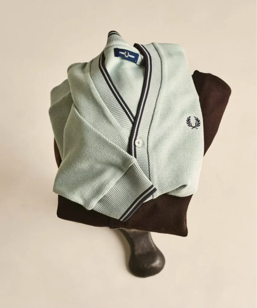 A pale mint-green Fred Perry cardigan with textured knit construction is draped over a dark brown cardigan on a sculptural wooden stand. Contrast striped ribbing along the collar, placket, and cuffs highlights the brand’s heritage-inspired design, while the embroidered Laurel Wreath logo adds a subtle signature detail