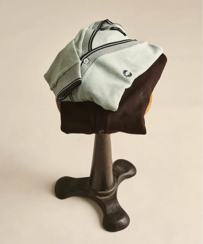 Two Fred Perry cardigans in complementary mint-green and deep brown colorways are layered together on a vintage-style wooden display stand. The textured knit fabric, ribbed trims, and embroidered Laurel Wreath logos emphasize the collection’s blend of classic British sportswear and contemporary style