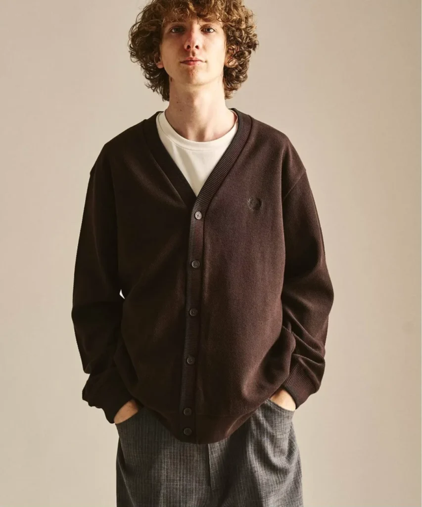 A model showcases a rich dark brown Fred Perry cardigan crafted from textured knit fabric with a clean V-neck silhouette and tonal embroidered Laurel Wreath logo. Paired with a white T-shirt and grey trousers, the cardigan delivers a refined, understated take on day layer