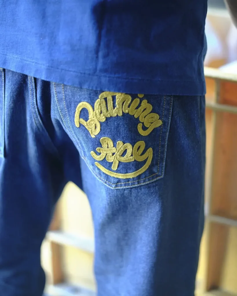 Close-up of A BATHING APE denim jeans with distressed ape-head pocket graphic and contrast stitching on dark indigo denim
