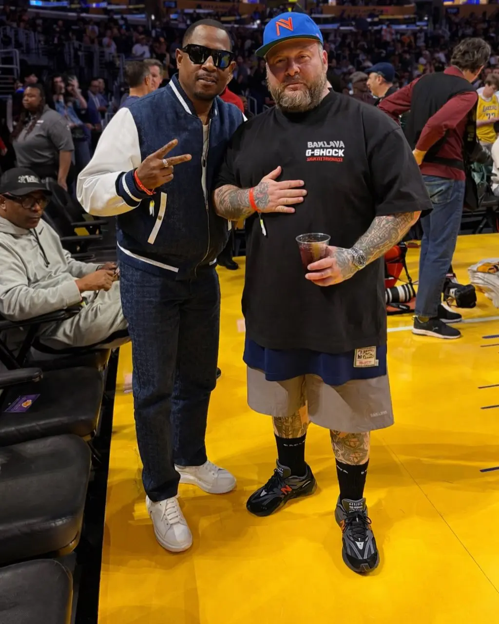 Courtside photo of two people posing at a basketball arena. One wears a navy varsity jacket with white sleeves and white sneakers, while the other sports a black Baklava x G-SHOCK T-shirt, layered shorts, a blue cap, and the Baklava x New Balance 2010 sneakers. The bright yellow hardwood court and spectators in the background create a lively game-day atmosphere