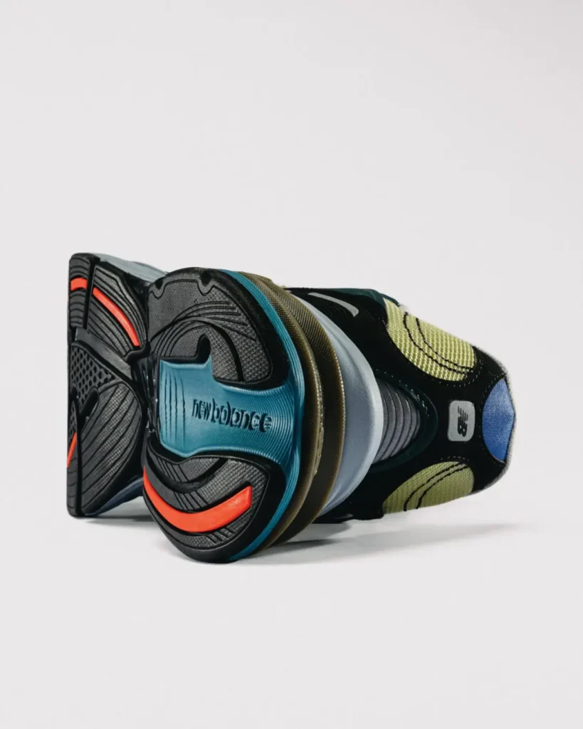 Studio product shot of the Baklava x New Balance 2010 viewed from the outsole and heel. The shoe features a segmented black rubber outsole with bright orange traction accents, a turquoise heel insert embossed with the New Balance logo, layered cushioning, and a mix of olive, gray, and black material