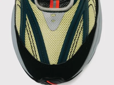 Close-up of the forefoot on the Baklava x New Balance 2010 showcasing pale olive mesh, dark teal structural overlays, black suede mudguards, reflective silver detailing, black rope laces, and a small red-orange pull tab at the base of the tongue. The image emphasizes the shoe's layered textures and technical construction