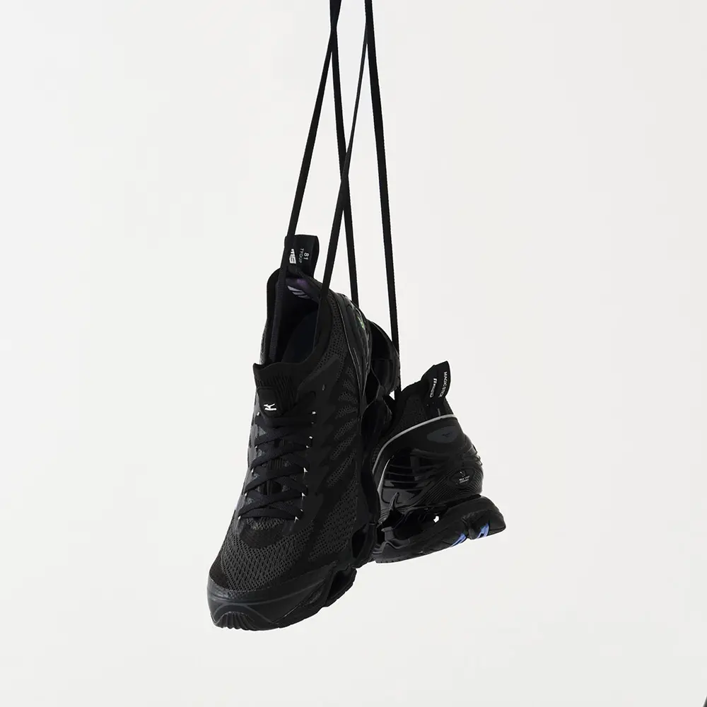 A pair of black Mizuno Wave Prophecy LS sneakers hangs from long black straps against a clean off-white studio backdrop. One shoe faces forward while the other reveals the sculpted heel and Wave sole, emphasizing the lightweight construction, breathable mesh upper, and premium connective style