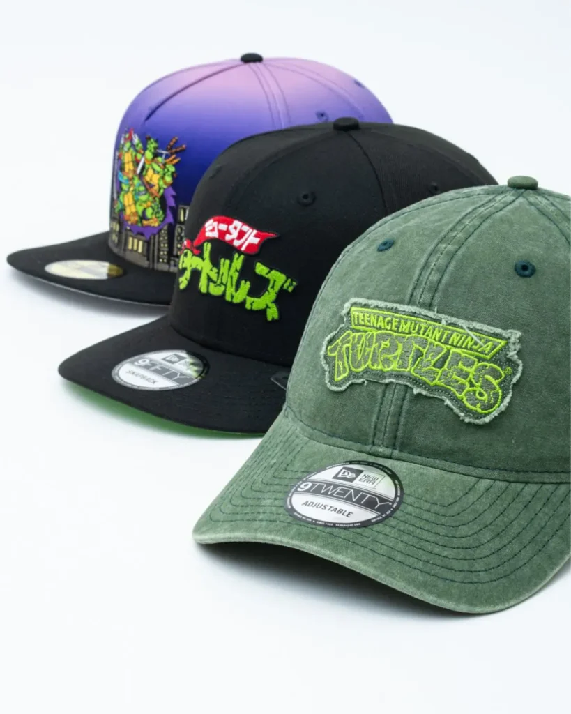 Close-up view of the Teenage Mutant Ninja Turtles x New Era headwear collection. In the foreground, a washed olive-green 9TWENTY adjustable cap showcases a frayed TMNT logo patch in neon green. Behind it sit a black 9FIFTY snapback with Japanese graphic embroidery and a purple gradient fitted cap featuring colorful turtle artwork and detailed cityscape stitching around the crown