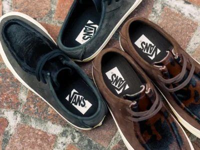 Overhead view of two Vans low-top moc-toe sneakers arranged on a brick-paved surface. One pair features black hairy suede panels with dark leather overlays, while the other combines brown leather with leopard-print pony hair accents, both finished with off-white mid