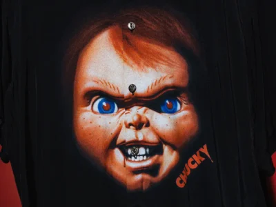 Close-up of a black Schott NYC camp-collar shirt featuring a large vintage-style graphic of Chucky’s menacing face across the front, accented by vivid blue eyes and orange lettering against a dark backdrop
